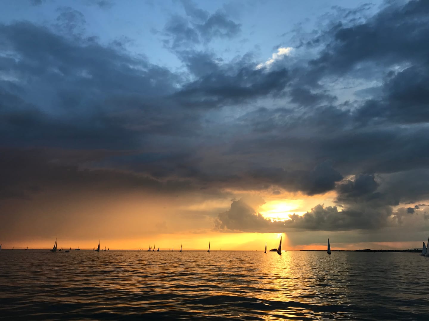 Gewitterwolken und Sonnenuntergang über dem Bodensee, am Horizont Segelboote.